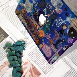 May include: A blue laptop cover with a collage design featuring characters and elements from a fantasy story. The cover includes the Apple logo and text that reads "Be careful what you wish for."