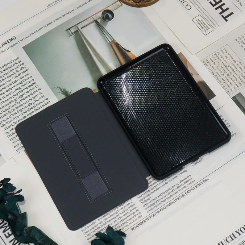May include: A black tablet case with a textured finish and a dark grey interior. The case is open, showing the tablet screen. The case has a built-in stand for hands-free viewing. The background includes newspaper cuttings.