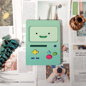May include: A mint green handheld gaming device with a smiling face on the screen. It features a yellow plus-shaped button, a blue triangle, and a pink circle. The device is set against a backdrop of newspaper clippings.