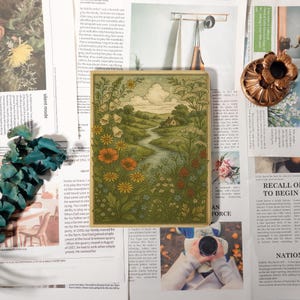 May include: A framed landscape illustration with a river, flowers, and a house. The artwork features a green and gold border and is surrounded by newspaper clippings and decorative elements. The illustration measures approximately 15 cm by 15 cm.