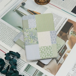 May include: A patchwork-style notebook with a variety of green and blue patterned fabrics. The notebook is rectangular and appears to be closed. The background includes newspaper clippings and a decorative object.