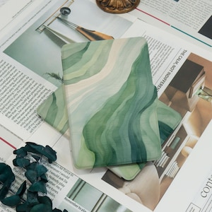 Könnte beinhalten: Eine rechteckige Handyhülle mit einem geschichteten Aquarelldesign in Grün- und Cremetönen. Die Hülle liegt auf einer Oberfläche mit Zeitungsausschnitten. Das Design hat ein fließendes, organisches Muster.