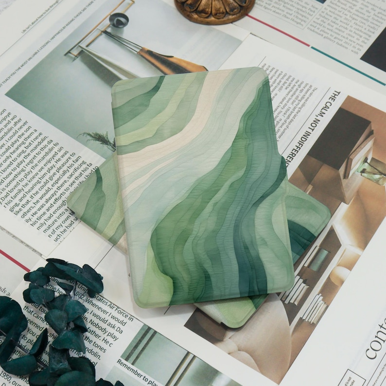 K&ouml;nnte beinhalten: Eine rechteckige Handyh&uuml;lle mit einem geschichteten Aquarelldesign in Gr&uuml;n- und Cremet&ouml;nen. Die H&uuml;lle liegt auf einer Oberfl&auml;che mit Zeitungsausschnitten. Das Design hat ein flie&szlig;endes, organisches Muster.