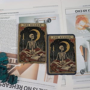 May include: Two tarot cards labelled "THE READER" are shown. Each card depicts a skeleton meditating, a crescent moon, and a candle. The cards have dark backgrounds with gold and red details, and are placed on a newspaper.