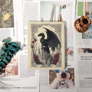 May include: A decorative, rectangular journal featuring a black dragon perched atop a castle, surrounded by red roses. The image has a vintage, gothic aesthetic. The journal is set against a backdrop of newspaper clippings and other decorative elements.