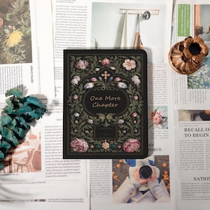 Peut inclure: Un journal noir avec des motifs floraux et une croix, portant l'inscription "One More Chapter". La couverture est ornée de fleurs roses, blanches et vertes. Le journal est entouré de coupures de journaux et d'éléments décoratifs.