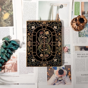 May include: A black decorative square with a gold floral design. The design features flowers and leaves within a gold frame. The square is surrounded by newspaper clippings and other decorative items.