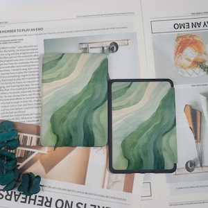K&ouml;nnte beinhalten: Zwei Tablet-H&uuml;llen mit einem wellenf&ouml;rmigen Muster in Gr&uuml;n und Wei&szlig;. Die rechteckigen H&uuml;llen haben einen schwarzen Rand. Das Design erinnert an Aquarellpinselstriche. Auf einer Zeitung pr&auml;sentiert.