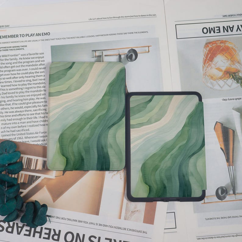 K&ouml;nnte beinhalten: Zwei Tablet-H&uuml;llen mit einem wellenf&ouml;rmigen Muster in Gr&uuml;n und Wei&szlig;. Die rechteckigen H&uuml;llen haben einen schwarzen Rand. Das Design erinnert an Aquarellpinselstriche. Auf einer Zeitung pr&auml;sentiert.
