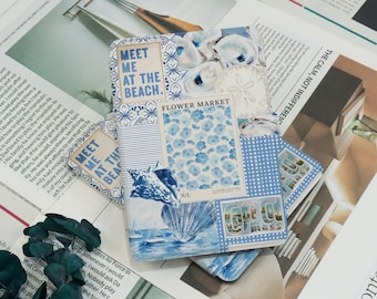 May include: A collection of decorative notebooks and cards with a coastal theme. The designs feature blue and white patterns, floral motifs, and seashell illustrations. Some items have text that reads "Meet Me at the Beach" and "Flower Market".
