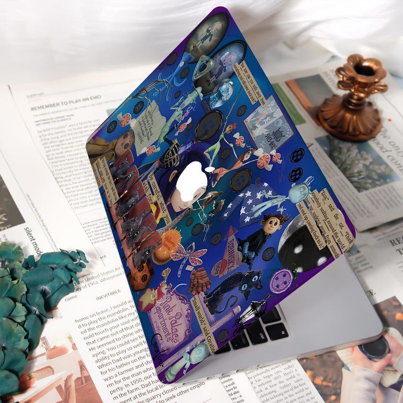 May include: A blue laptop case with a collage design featuring illustrations and text from the movie "Coraline." The case has a white Apple logo cutout and a purple border. The design includes images of buttons, a black cat, and the text "Pink Palace apartments."