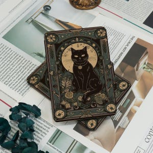 May include: Two tarot cards featuring a black cat illustration. The cat sits in front of a full moon, surrounded by floral and insect motifs. The cards have a dark teal border with gold accents, and are placed on a newspaper background.