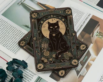 Könnte beinhalten: Zwei Tarotkarten mit einer Illustration einer schwarzen Katze. Die Katze sitzt vor einem Vollmond, umgeben von floralen und Insektenmotiven. Die Karten haben einen dunkelblauen Rand mit goldenen Akzenten, auf einem Zeitungs-Hintergrund.