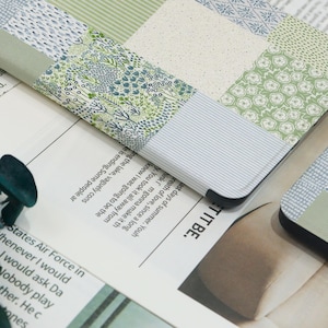 May include: A close-up of a patchwork-style laptop case with a variety of green and blue floral and patterned fabrics. The case is resting on a newspaper with text. The edges of the case are black.
