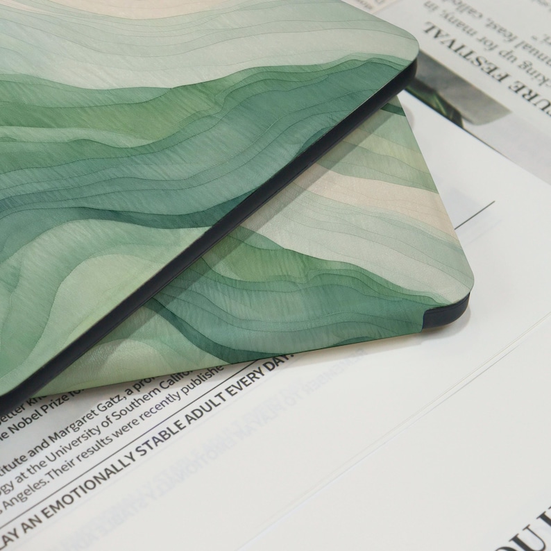 K&ouml;nnte beinhalten: Nahaufnahme einer Handyh&uuml;lle mit einem geschichteten Aquarelldesign in Gr&uuml;n- und Beiget&ouml;nen. Die H&uuml;lle hat einen schwarzen Rand und eine glatte Oberfl&auml;che. Das Design &auml;hnelt H&uuml;geln oder abstrakten Wellen.