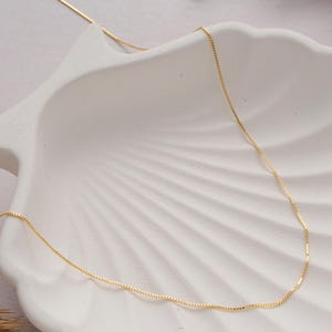 May include: A delicate gold chain necklace, displayed on a white seashell-shaped surface. The chain is thin and has a box link pattern.