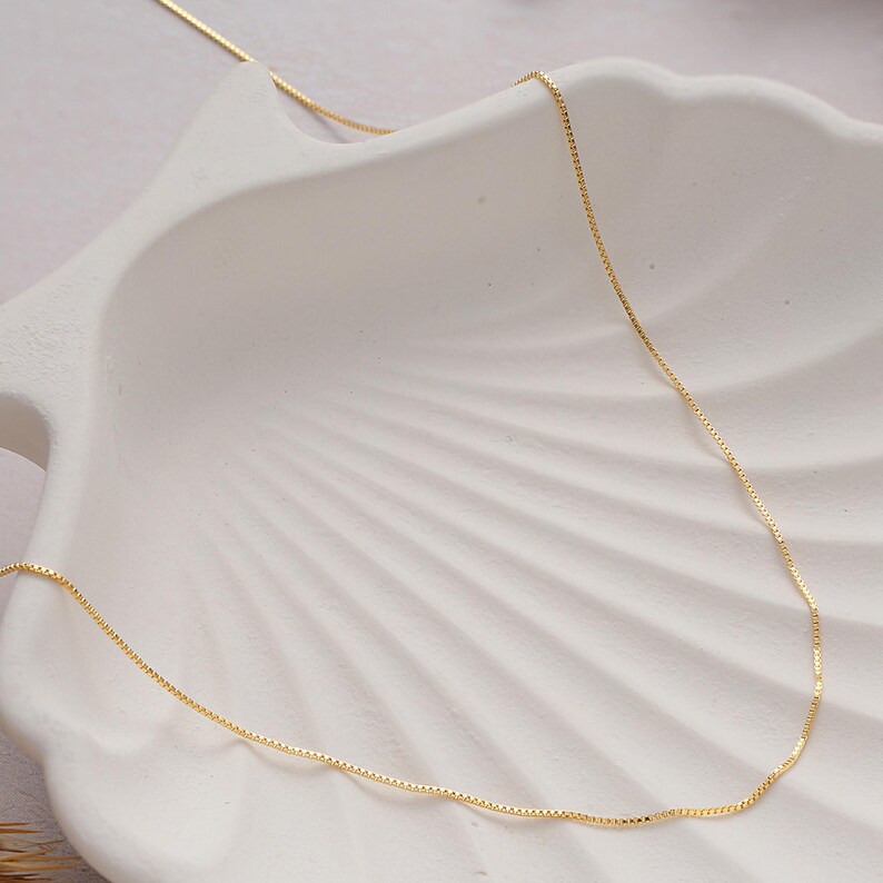 May include: A delicate gold chain necklace, displayed on a white seashell-shaped surface. The chain is thin and has a box link pattern.