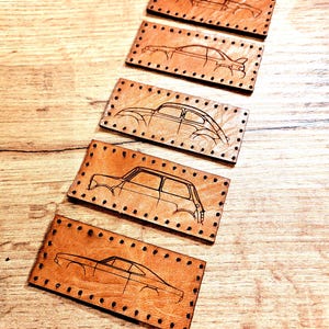 May include: Five leather keychains with different car silhouettes engraved on them. The keychains are brown and have a stitched border.