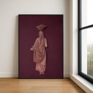 Op de afbeelding: Een ingelijst kunstwerk dat een vrouw afbeeldt die een mand op haar hoofd en een kruik draagt. Het reliëf is in roze en bruine tinten tegen een diepe bordeauxrode achtergrond. De vrouw is gehuld in stof en het kunstwerk is in een zwart frame.