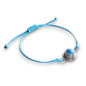 Könnte beinhalten: Ein blaues Armband aus Schnur mit einem silbernen Anhänger, der eine klare Glaskuppel mit blauem und weißem Glitzer enthält.