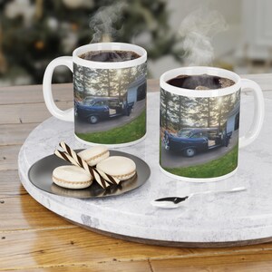 Puede incluir: Dos tazas de café de cerámica blanca con una imagen de un coche azul clásico aparcado frente a un garaje. Las tazas están llenas de café caliente, y el vapor se eleva desde la parte superior. Un plato con macarons y una cuchara están en una bandeja de mármol.