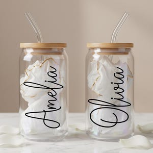May include: Two clear glass tumblers with bamboo lids and bent glass straws. Each tumbler features a custom name in black cursive script, with the names Amelia and Olivia printed on the front. The glasses are filled with white tissue paper.
