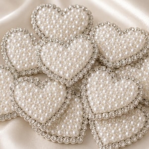 Peut inclure: Une collection d'ornements en forme de cœur avec un centre rempli de petites perles blanches artificielles et une bordure composée de strass transparents étincelants. Chaque cœur est uniforme, présentant un aspect texturé et superposé.