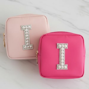 May include: Two square cosmetic bags, one light pink and one hot pink, with gold zippers. Each bag features a large letter "I" embellished with pearls and rhinestones, creating a glamorous and personalized accessory.