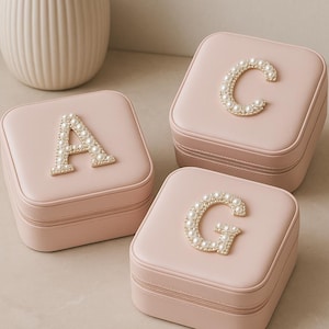 May include: Three pink square jewelry boxes with gold zippers. Each box has a different letter, A, C, and G, made of small pearls. A white vase is in the background.