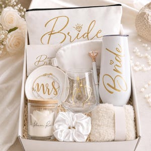 May include: A gift box containing a white canvas pouch with gold Bride text, a white sleep mask, a white pen with a diamond topper, a white tumbler with gold Bride text, a small dish with Mrs text, a pearl bracelet, a glass, a candle, a scrunchie, and socks.