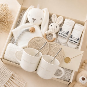 May include: A gift box containing a white plush bunny security blanket, a wooden pacifier clip with grey beads, a crocheted bunny rattle, two pairs of white baby socks with embroidered faces, two speckled mugs labeled mama bear and papa bear, and two round woven coasters.