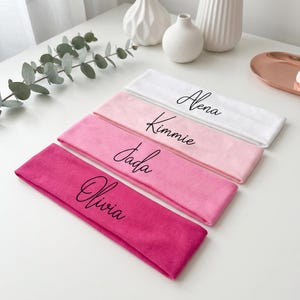 May include: Four headbands in shades of pink and white are arranged on a white surface. Each headband is personalized with a name in black script: Alena, Kimmie, Jada, and Olivia. Decorative vases and greenery are in the background.
