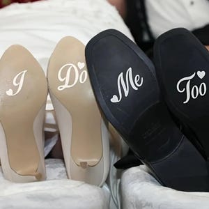Puede incluir: Zapatos de boda con las palabras "I Do" impresas en las suelas de los tacones beige y "Me Too" en las suelas de los zapatos negros. El texto está en una fuente blanca con un símbolo de corazón.