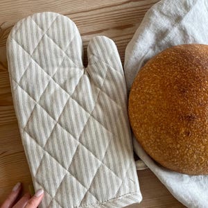 May include: A beige and white striped oven glove with a quilted diamond pattern sits beside a round loaf of bread on a linen cloth. The bread has a golden-brown crust. The items are on a light-coloured wooden surface.