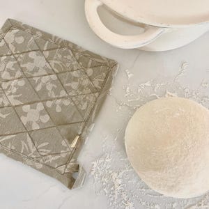 May include: A beige floral-patterned quilted oven glove sits next to a white ceramic pot with handles and a ball of dough dusted with flour. The glove has a loop for hanging.