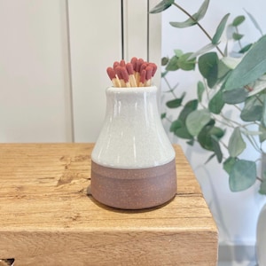 May include: A white ceramic match holder with a brown base, filled with red-tipped matches.