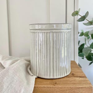 May include: A white ceramic planter with vertical ridges. The planter has a smooth, glazed finish and a slightly rounded shape.