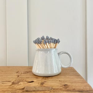 May include: A white ceramic match holder with a handle, filled with grey-tipped matches.