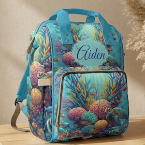 Può includere: Uno zaino turchese con un colorato design a barriera corallina, con tartarughe marine e pesci. Lo zaino ha una maniglia superiore, spallacci regolabili e una tasca frontale. Il nome "Aiden" è stampato su un ovale blu.