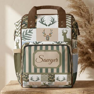 May include: A personalized backpack with a brown handle and trim. The front features the name "Sawyer" in a decorative font, set against a striped background. The design incorporates deer silhouettes, antlers, and camouflage patterns in shades of green, brown, and white.