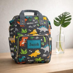May include: A grey dinosaur-themed backpack with a teal name patch that reads "Jacob." The backpack features a repeating pattern of colourful dinosaurs, palm trees, and the word "ROARRR." It has a grey handle and straps, and gold-coloured zips.