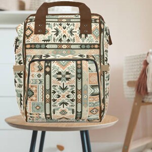 May include: A patterned backpack with a geometric design in shades of beige, brown, and teal. The bag has a top handle and adjustable straps. It features multiple pockets and a front flap. The backpack is sitting on a small wooden table.
