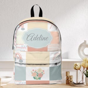 Könnte beinhalten: Ein pastellfarbener Rucksack mit einem Patchwork-Design mit Regenbogen, Blumen und Wolken. Der Rucksack hat einen schwarzen Griff und Träger sowie ein hellblaues Oval mit dem Namen "Adeline" in Schreibschrift.