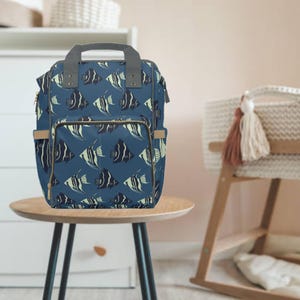 May include: A blue diaper bag featuring a repeating pattern of black and white fish. The bag has a grey handle and tan accents. It is placed on a small, round wooden table.