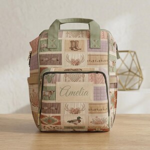 May include: A sage green and beige quilted backpack featuring a patchwork pattern of western-themed illustrations including cowboy boots, longhorn cattle, antlers, cacti, and a duck. A rectangular patch on the front pocket displays the name Amelia.