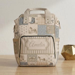 May include: A patchwork diaper bag with a top handle and multiple pockets. The bag features a variety of patterns, including horses, boots, and bows, in shades of beige, blue, and cream. The name "Amelia" is displayed on the front.