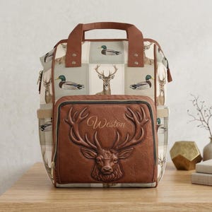 May include: A backpack featuring a tan and cream grid pattern with illustrations of deer and mallard ducks. The front pocket is made of brown faux leather with an embossed deer head and the name Weston printed in a cursive script font.