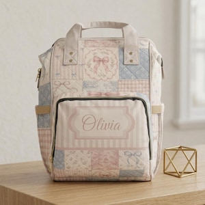 Puede incluir: Una mochila acolchada con un diseño de patchwork en tonos suaves de rosa, azul y crema. El patrón incluye ilustraciones de lazos y motivos florales. Un bolsillo frontal muestra una etiqueta rectangular con el nombre Olivia impreso en cursiva.