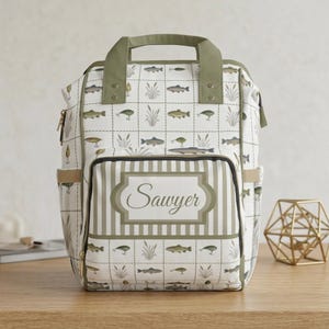 May include: A white backpack with a fishing theme, featuring fish and aquatic plants. The bag has a green handle and trim, with the name "Sawyer" on the front. It has multiple pockets and a striped front panel.