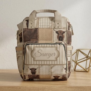 May include: A beige and brown patchwork backpack with a handle and shoulder straps. The design includes images of Highland cows, gingham, and striped patterns. The name "Sawyer" is displayed on the front.